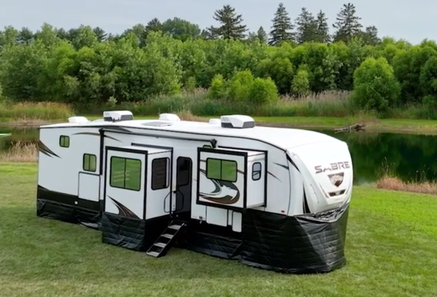 A fifth wheel RV with insulated RV skirting on grass next to a pond.