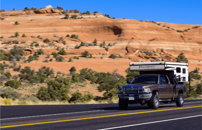 A pop up truck bed camper driving on a road against a red rock background.