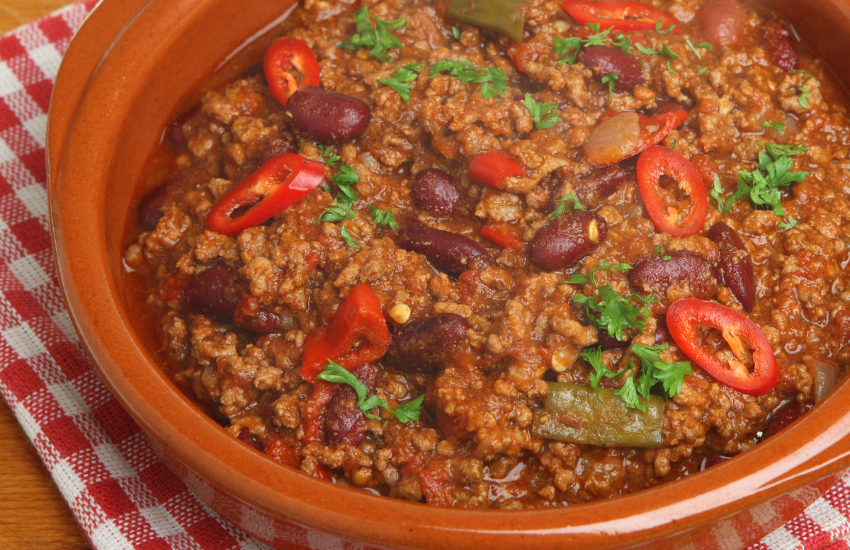 A red bowl on a chequered cloth with chilli con carne in it.